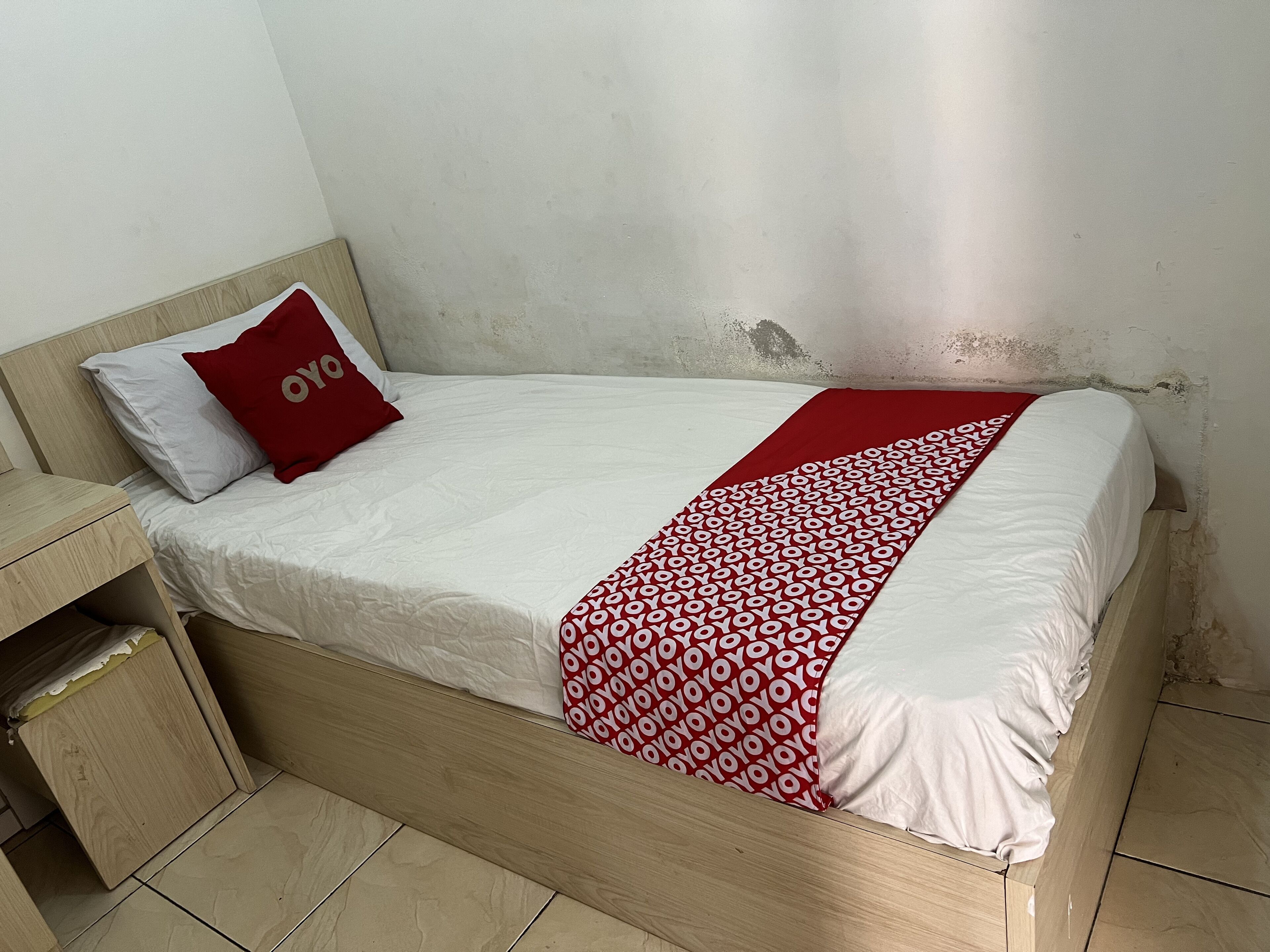Una cama individual con colchón blanco y manta de diseño rojo y blanco. Tiene un cabecero de madera y una mesita de noche pequeña a la izquierda. La pared es lisa y presenta marcas de uso.