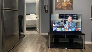 A living room with a television displaying a show, a refrigerator, a bed, and a painting on the wall. - Cozy Get-Away Suite (Regina)