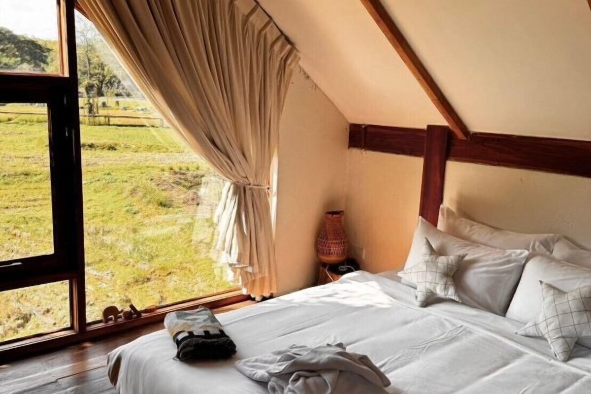 Un dormitorio con un ventanal grande, una cama con almohadas, una lámpara y suelo de madera.