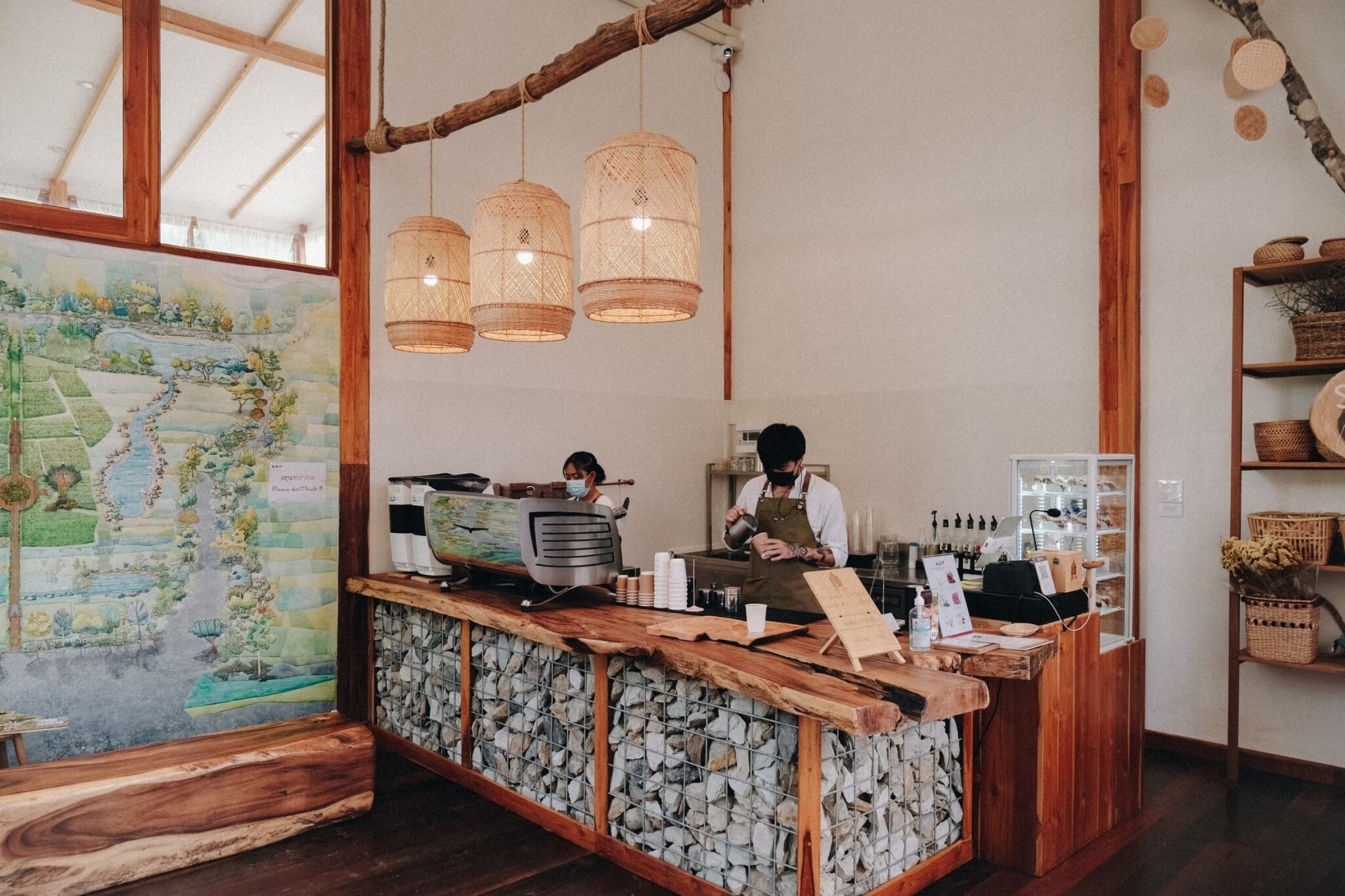 Interior de café rústico com balcão de madeira, luminárias de vime penduradas e uma grande pintura de paisagem na parede.