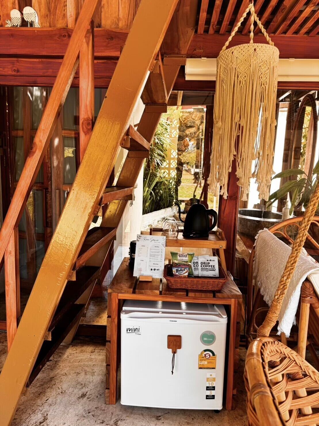 Une pièce chaleureuse avec un escalier en bois, un luminaire suspendu en macramé, un coin café avec un mini-réfrigérateur et des meubles en osier.