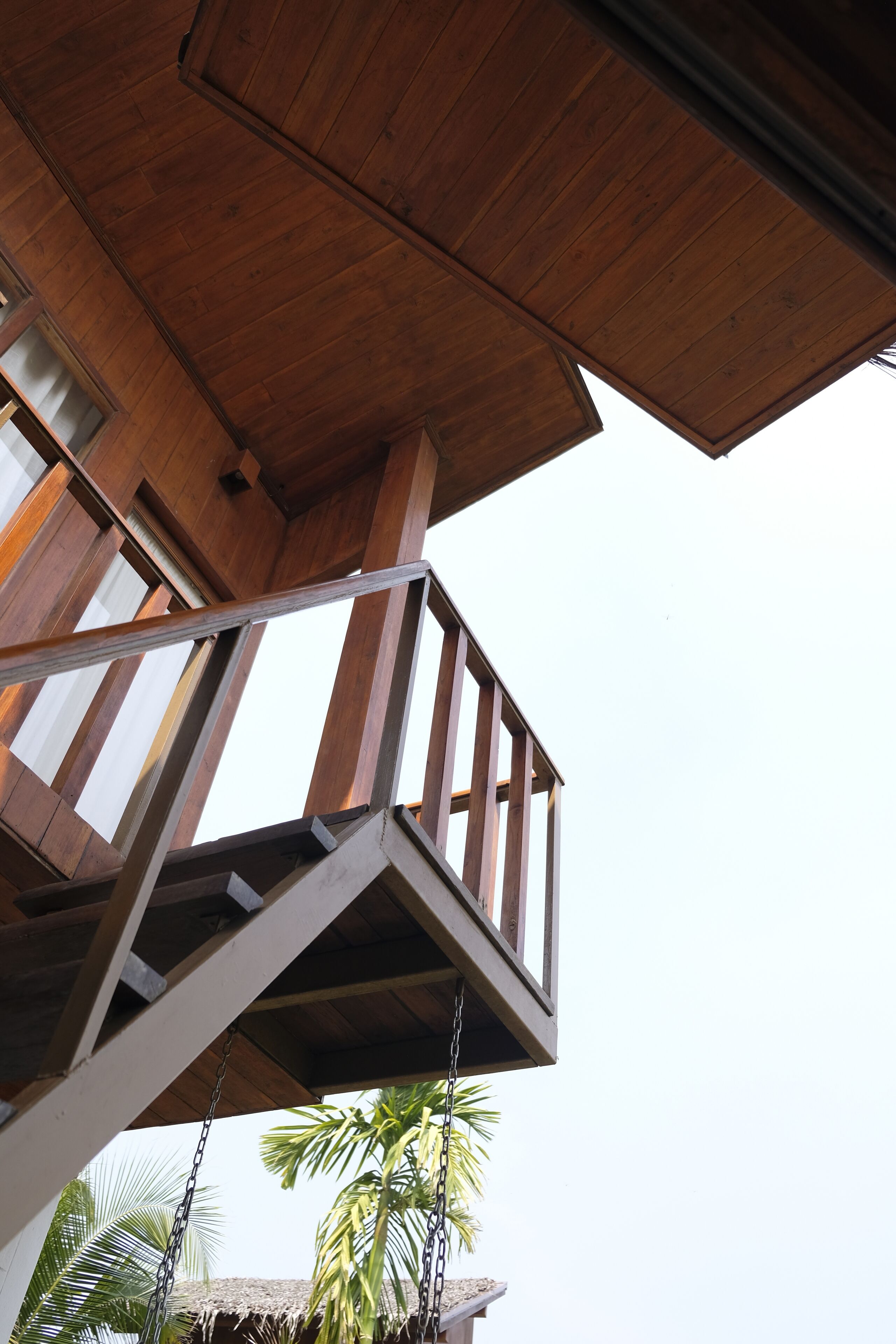 Un balcon en bois doté d’une rambarde en métal, offrant une vue sur une construction à toit de chaume et des palmiers.