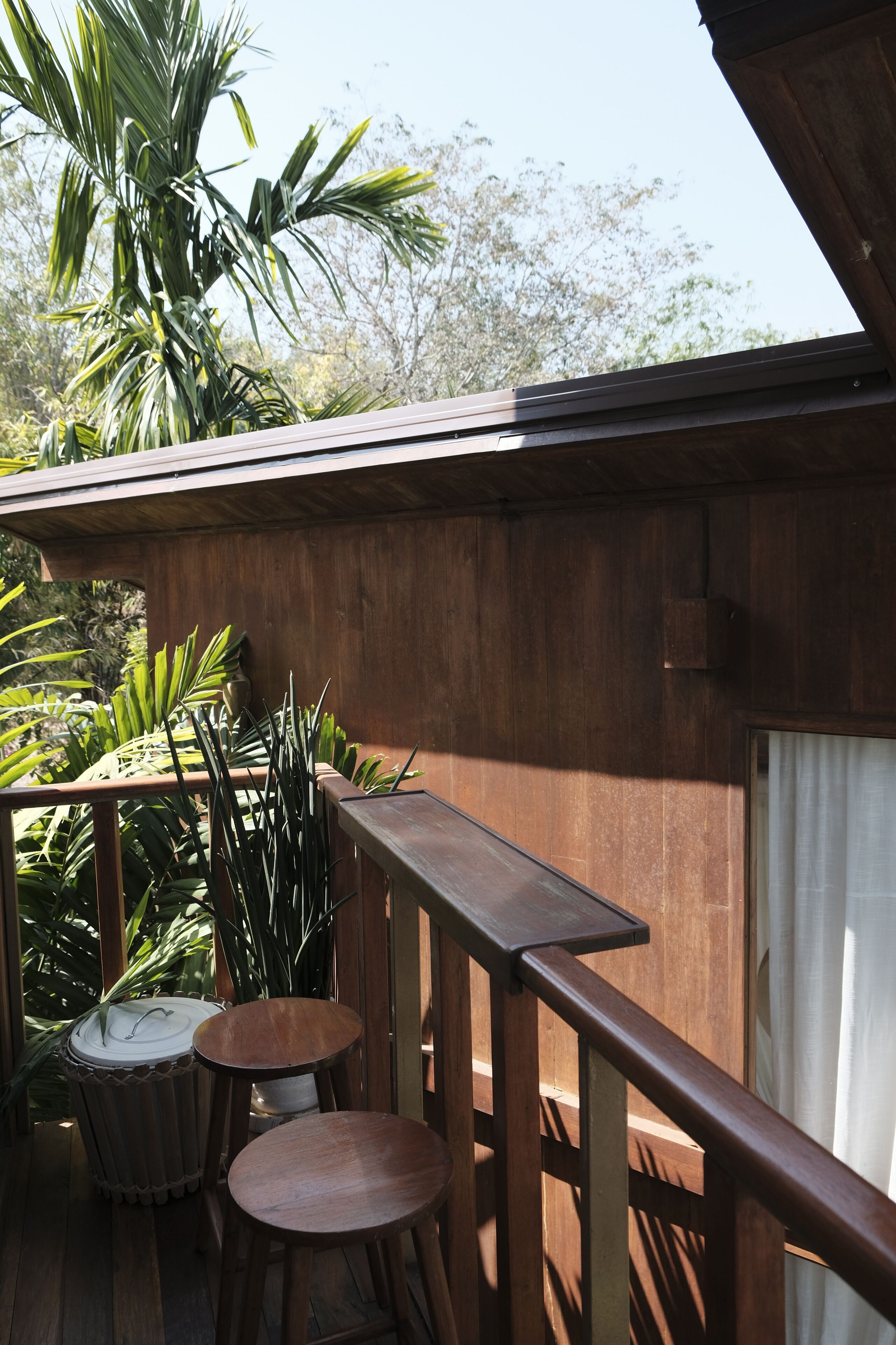 A wooden deck with two round wooden stools and a small table, surrounded by various plants and a large tree.