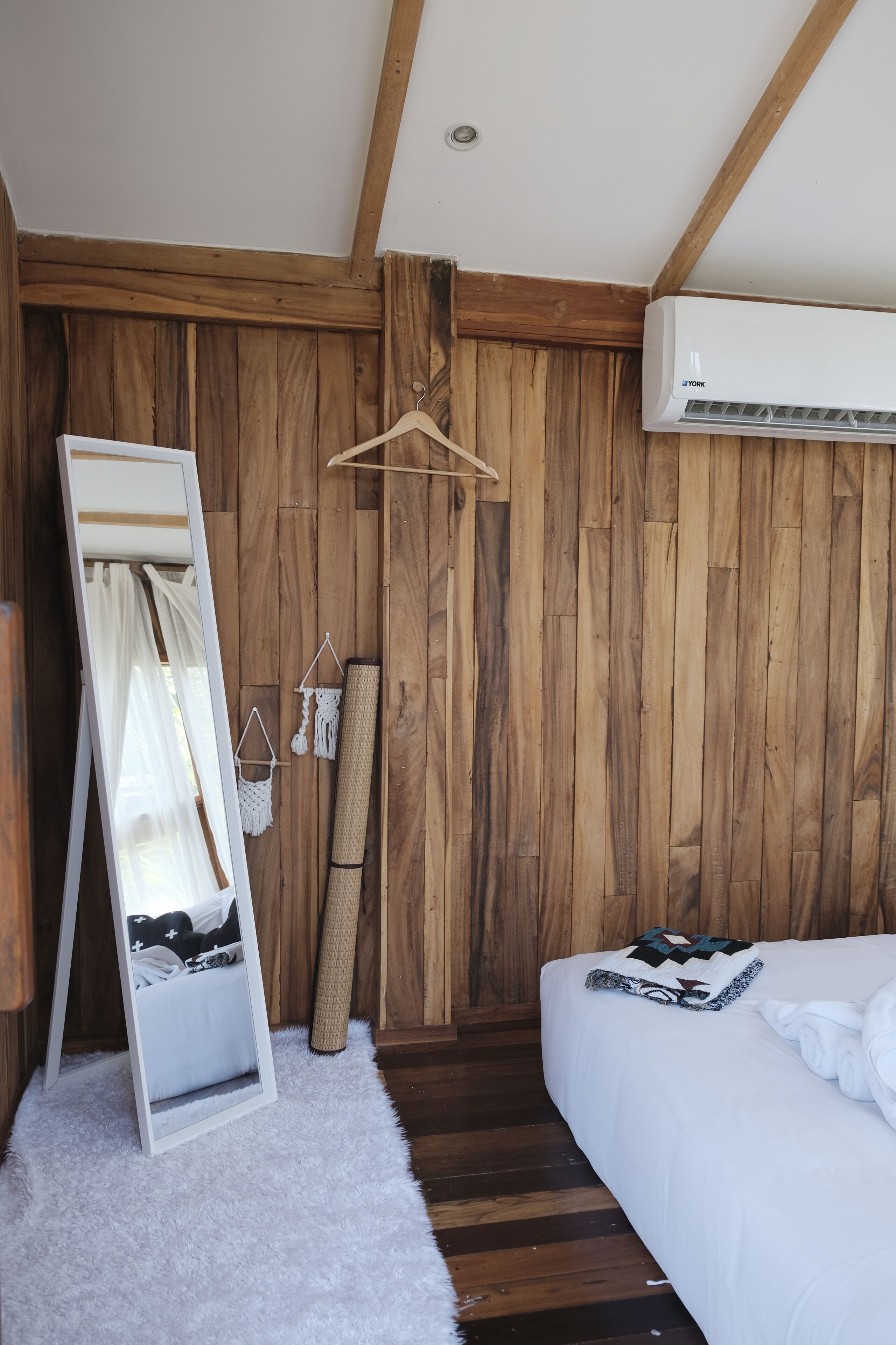 A bedroom with wooden walls, a white bed, a hanging mirror, and a white fluffy rug.