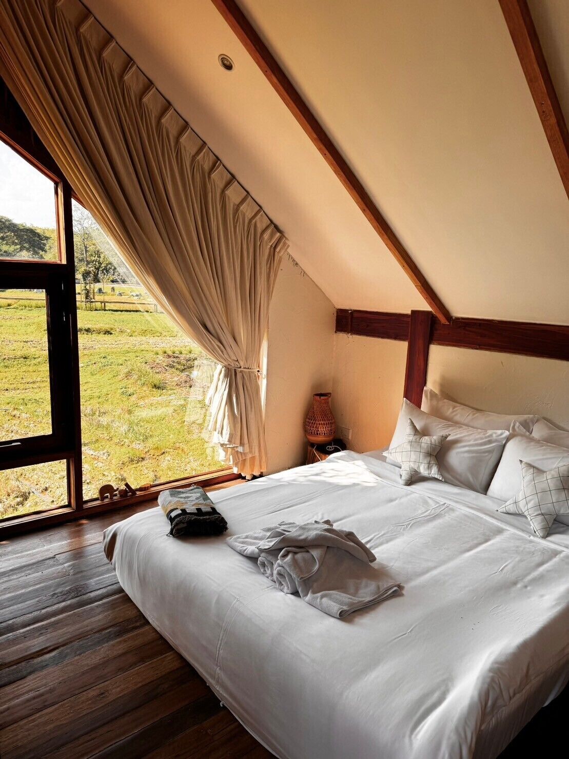 Een slaapkamer met een groot raam, houten balken, een bed met witte lakens en uitzicht op een grasveld.