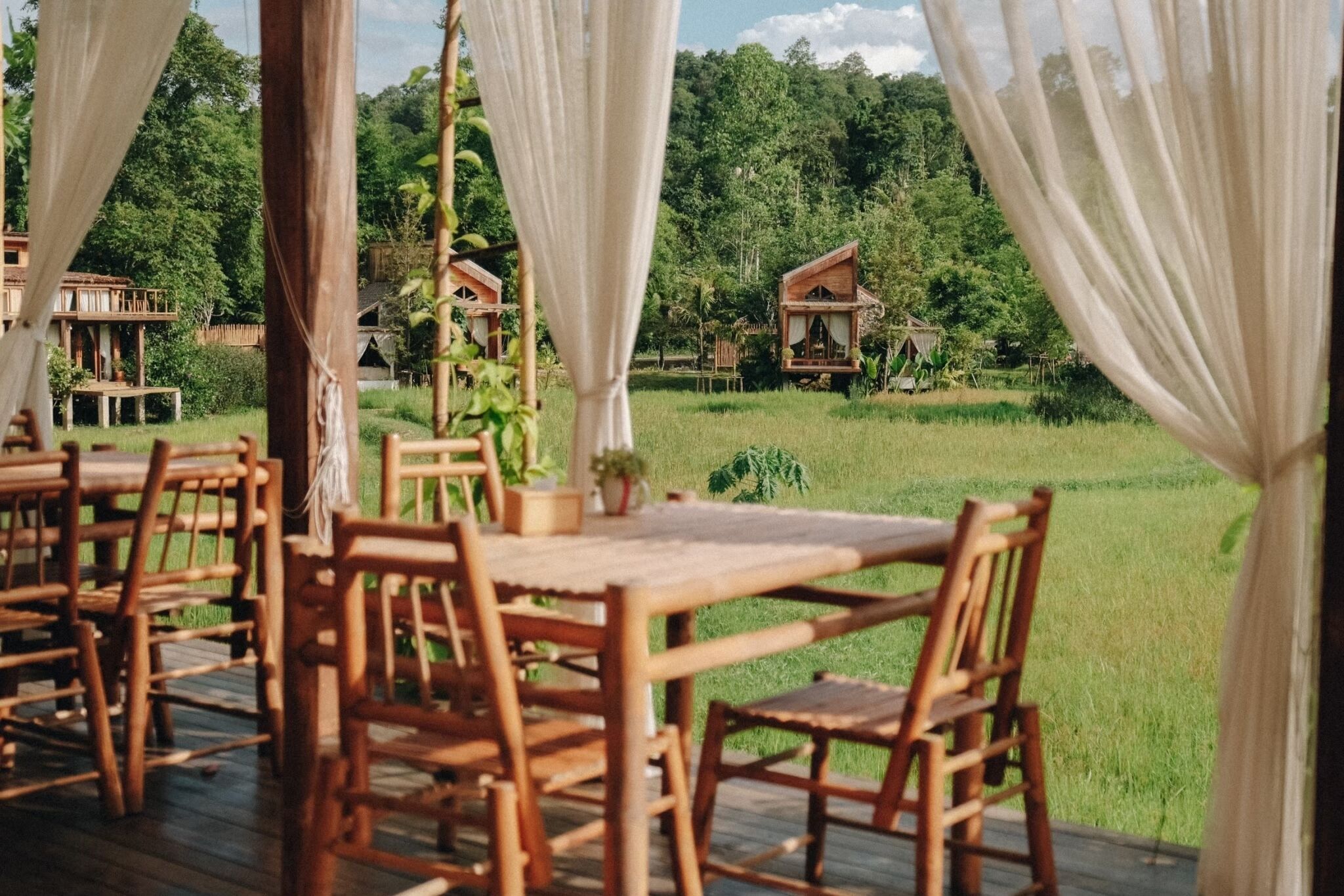 A wooden deck with a table and chairs, surrounded by white curtains, overlooking a lush green landscape with wooden cabins in the background.