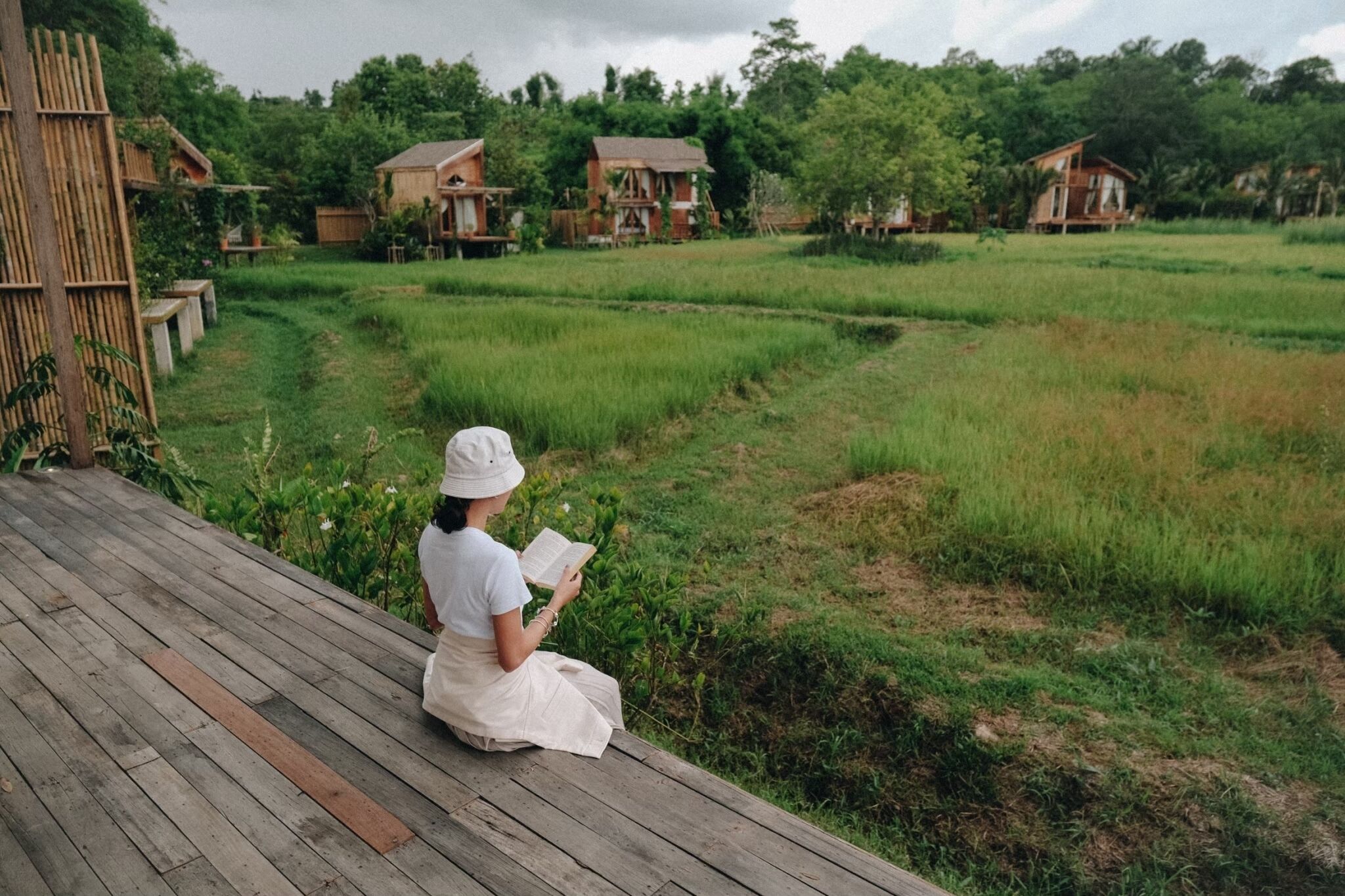 A person sitting on a wooden deck reading a book, overlooking a rice field with several wooden houses in the background.