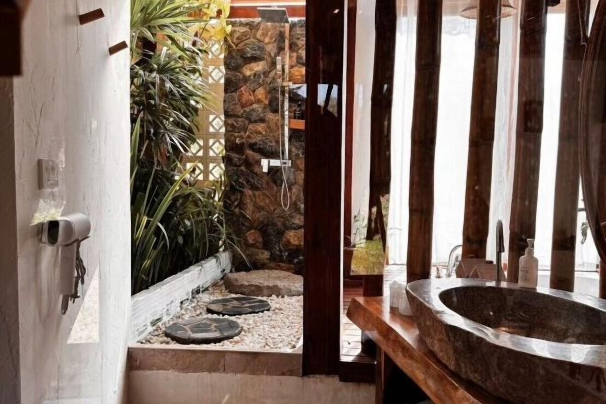 Un baño con lavamanos de piedra, zona de ducha y un pequeño jardín con plantas y pared de piedra.