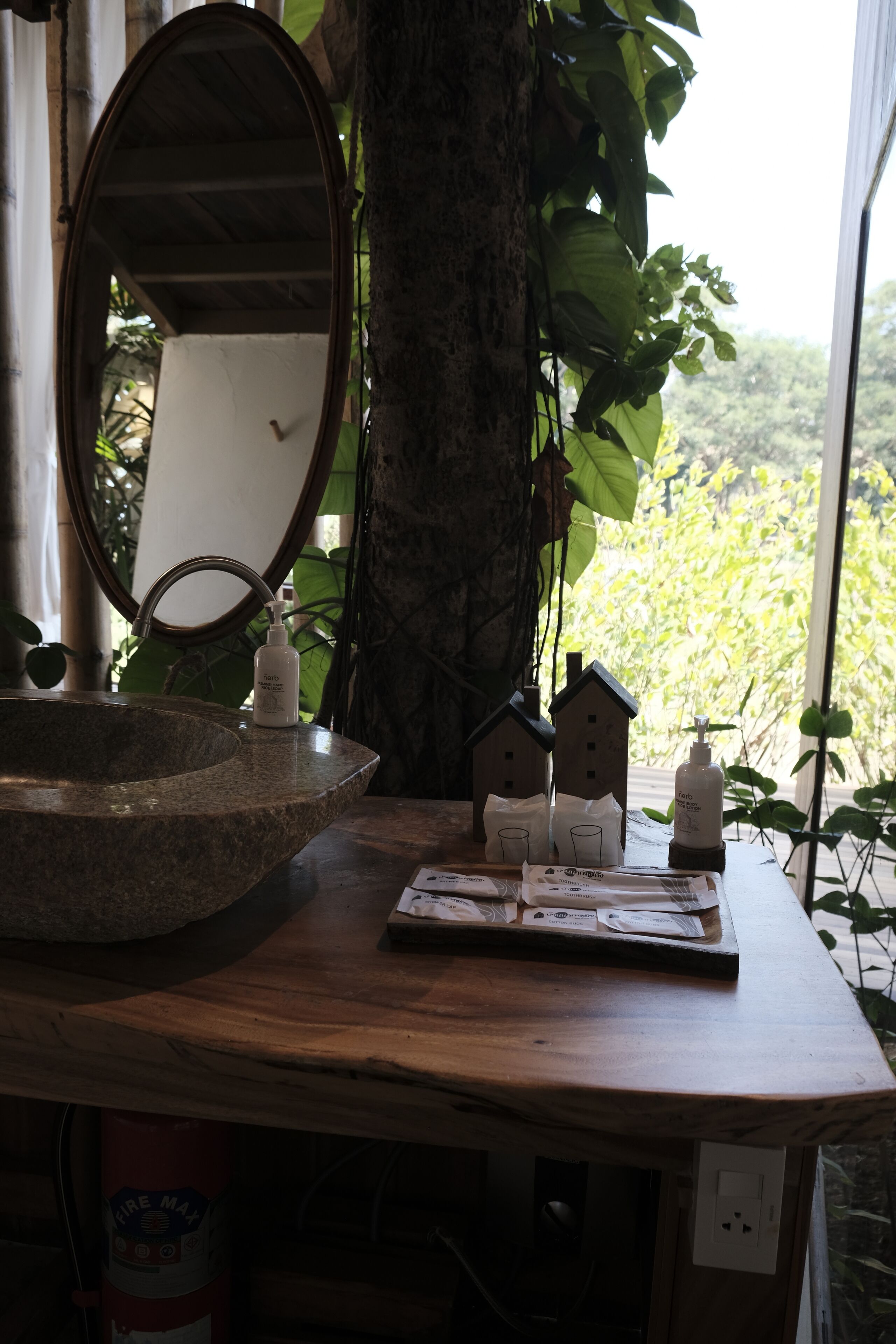 Une salle de bain avec un lavabo en pierre, un miroir, une petite table en bois sur laquelle sont posés des articles de toilette, et une vue sur la verdure à l’extérieur.