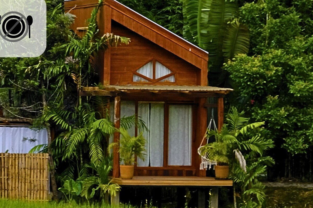 Una casa de madera rodeada de abundante vegetación, con un letrero que indica prohibido fumar, prohibido mascotas y no se permite comer fuera.