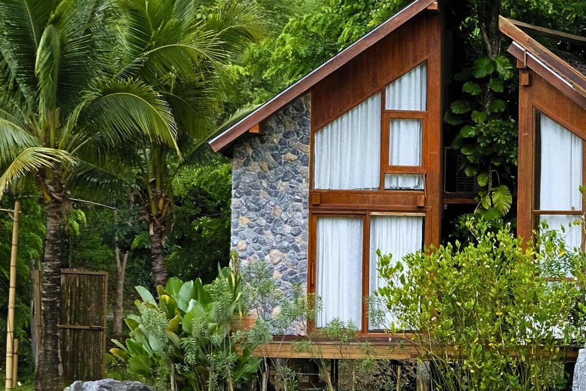 Una casa de madera rústica con paredes de piedra, rodeada de abundante vegetación y una gran roca al frente.