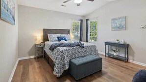A bedroom with a bed, bedside table, bench, ceiling fan, and a window with curtains. - Beach House Vacation  (Wilmington)