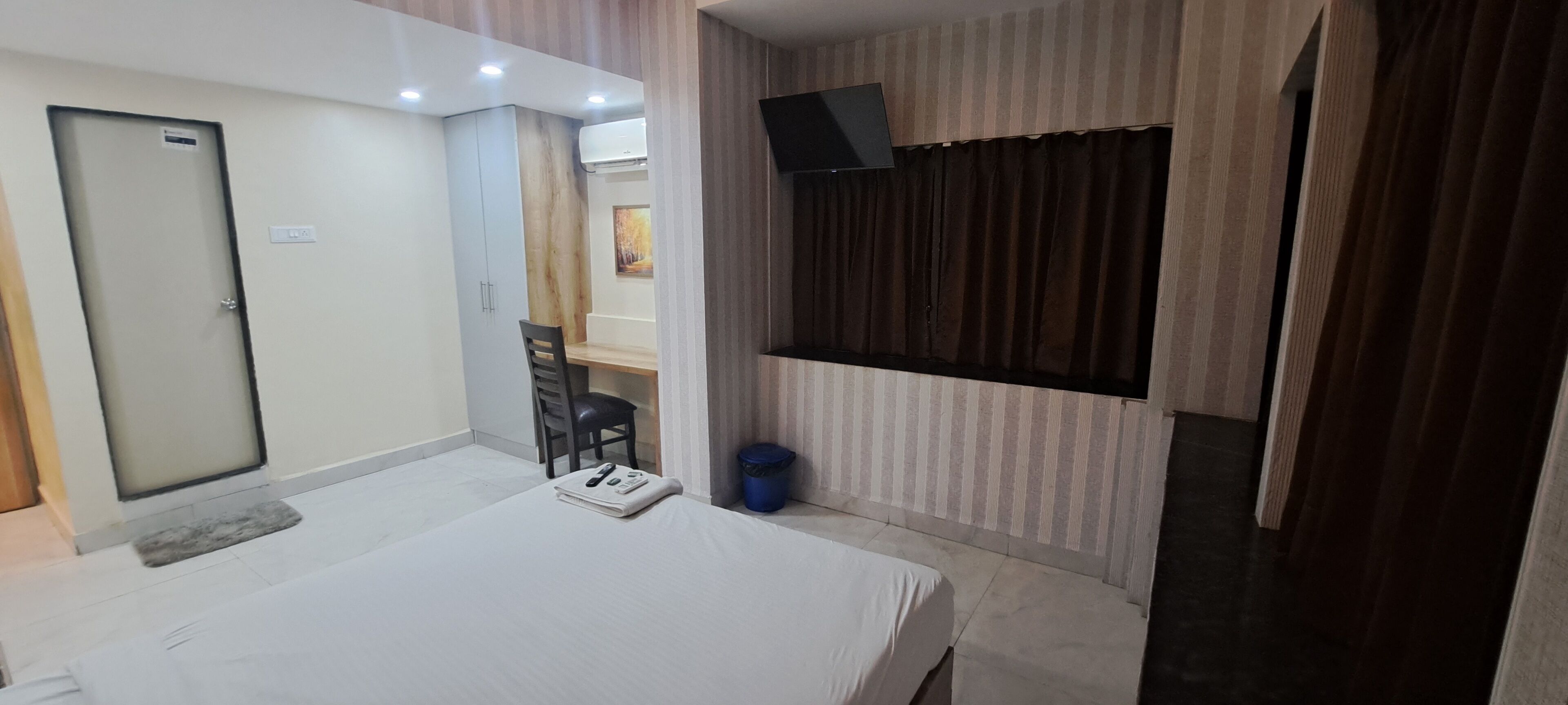 A hotel room with a bed, a desk, a chair, a TV mounted on the wall, and a window with curtains.