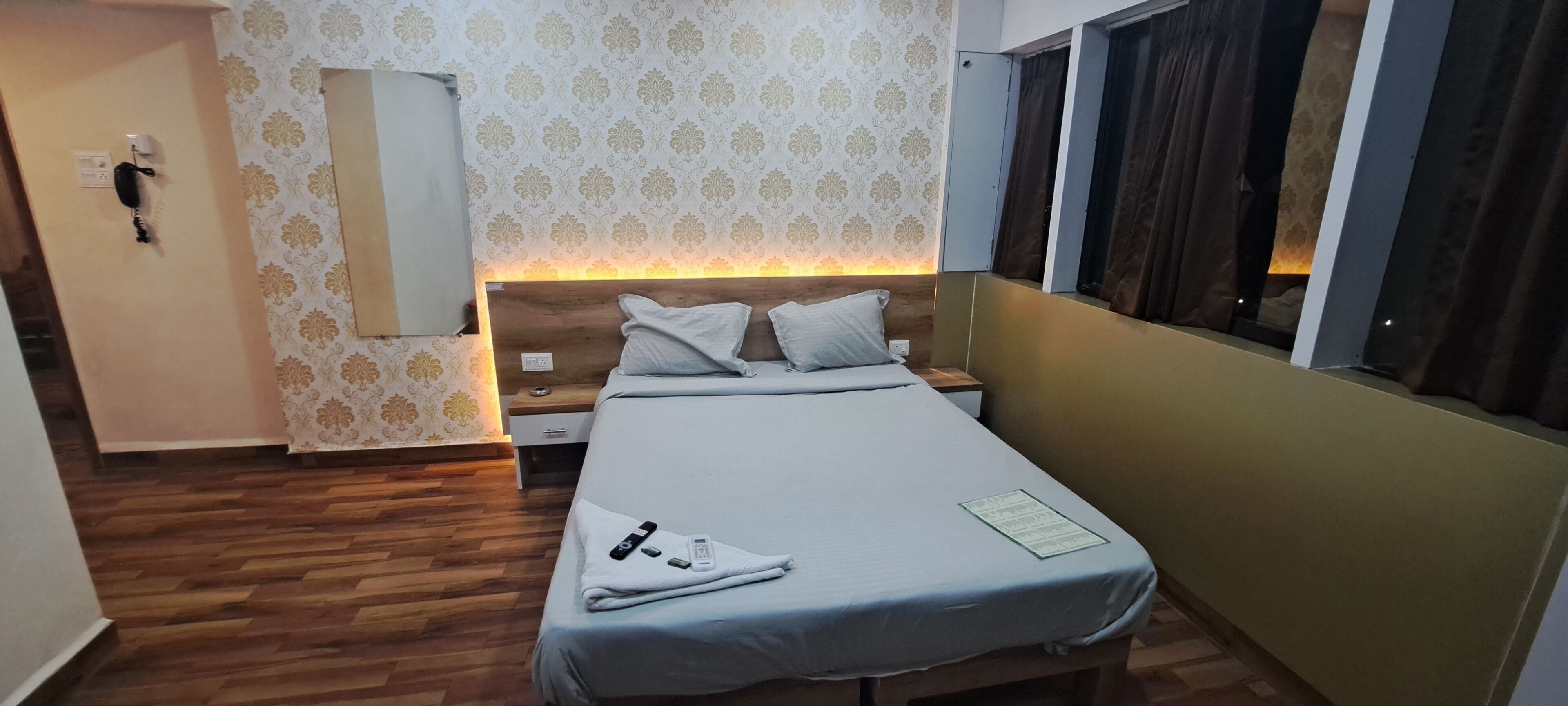 A neatly made bed with a grey bedspread and pillows, a bedside table with a lamp, and a wall-mounted phone.