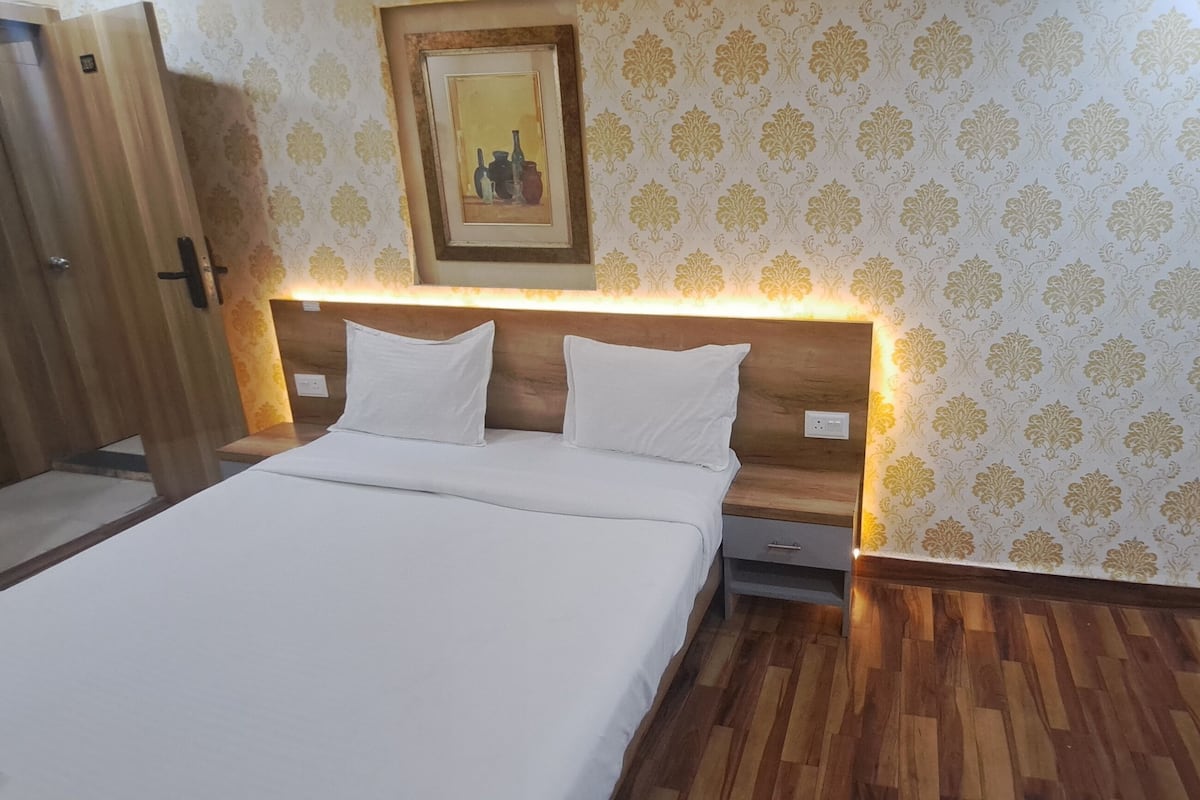 A hotel room with a wooden bed, white bedding, a nightstand, a wall-mounted lamp, and patterned wallpaper.
