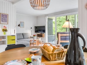 A modern living room with a sofa, TV, dining table, and a view of greenery outside.