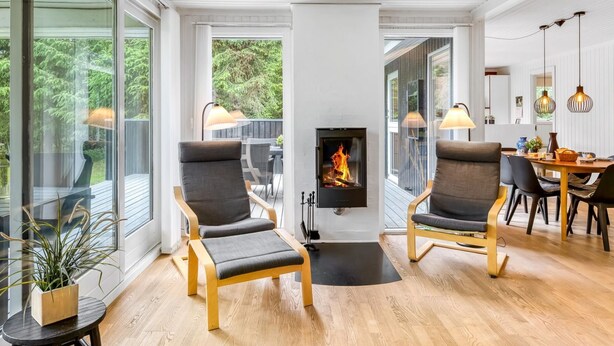 Un salon moderne avec une cheminée, deux fauteuils, un coin repas et de grandes fenêtres.