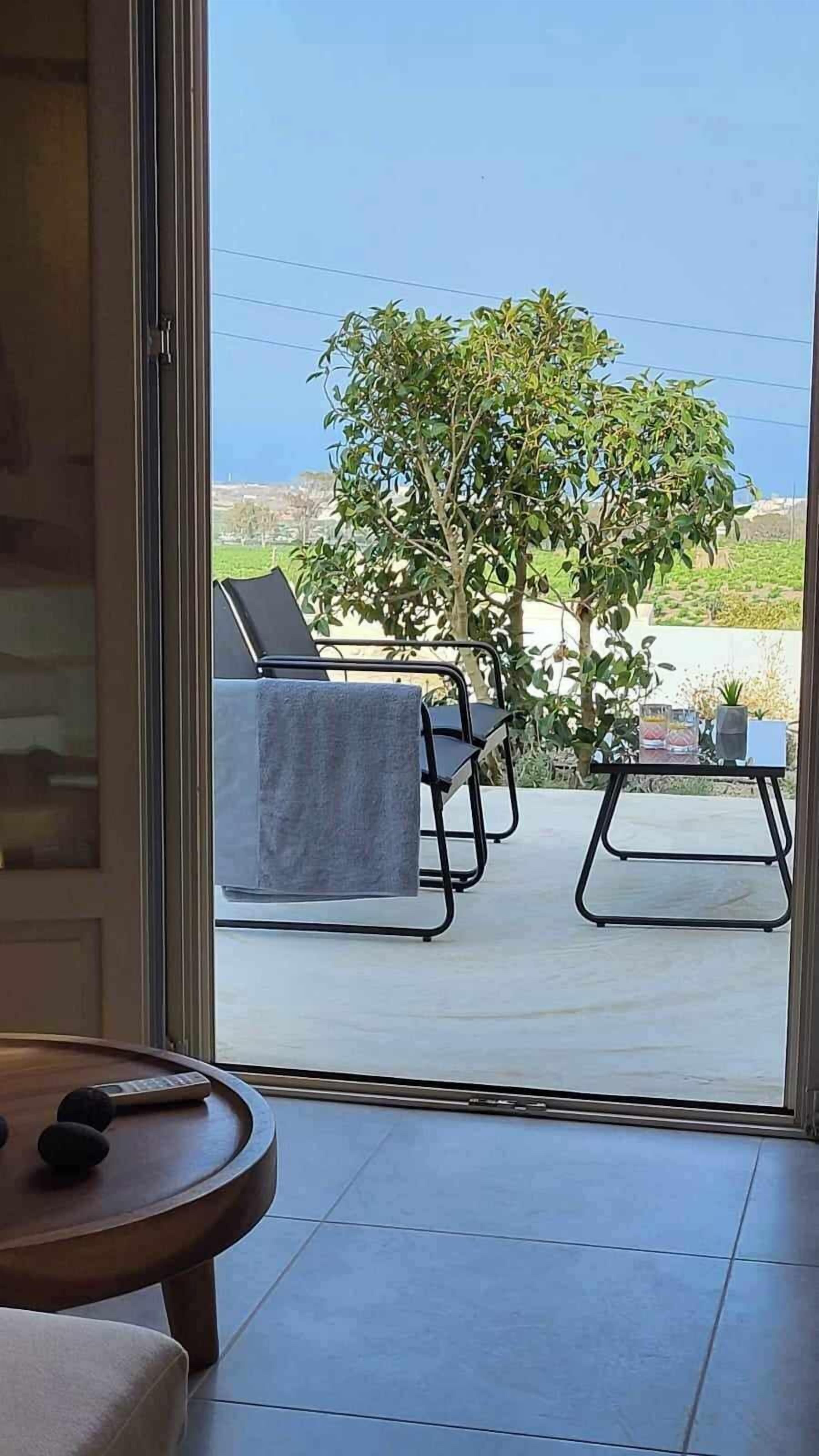 Habitación con puerta de vidrio corredera que da a un espacio exterior con dos sillas, una mesa, una planta pequeña en maceta y vistas a un árbol y el paisaje.