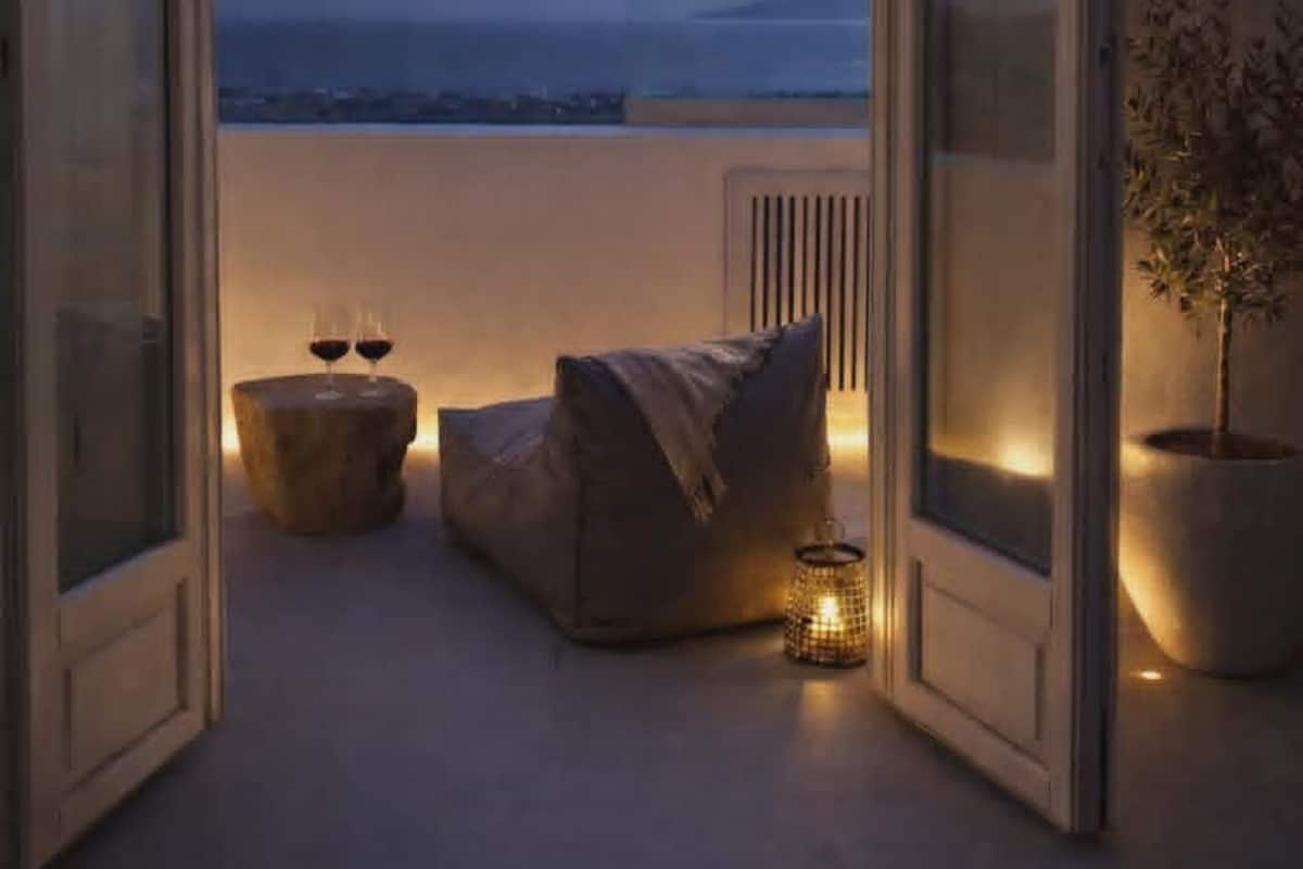 A room with a view of the sea, a bed, a small table with glasses, a chair, and a potted plant.