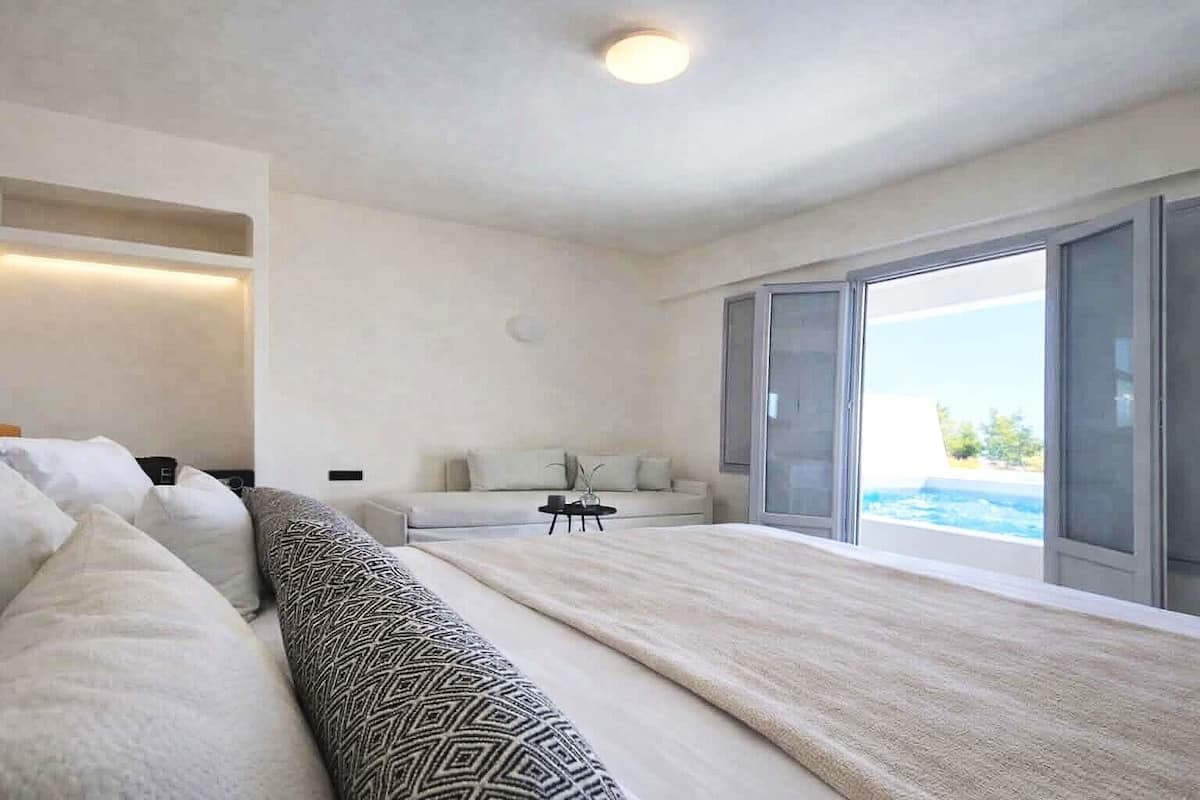 A bedroom with a large bed, a sofa, a small table, and a sliding glass door leading to a pool.