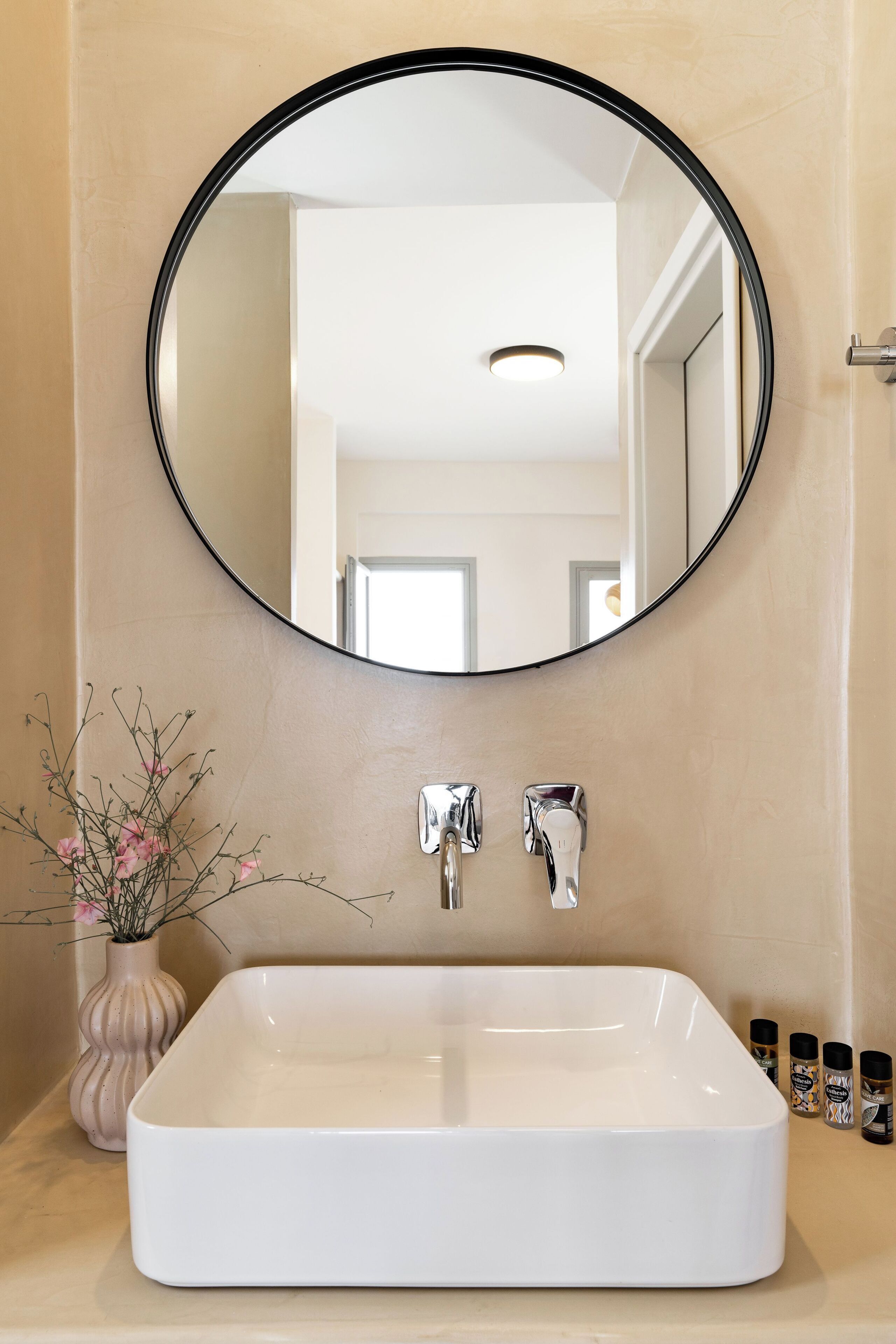 A bathroom with a white sink, a round mirror, and a vase with flowers.