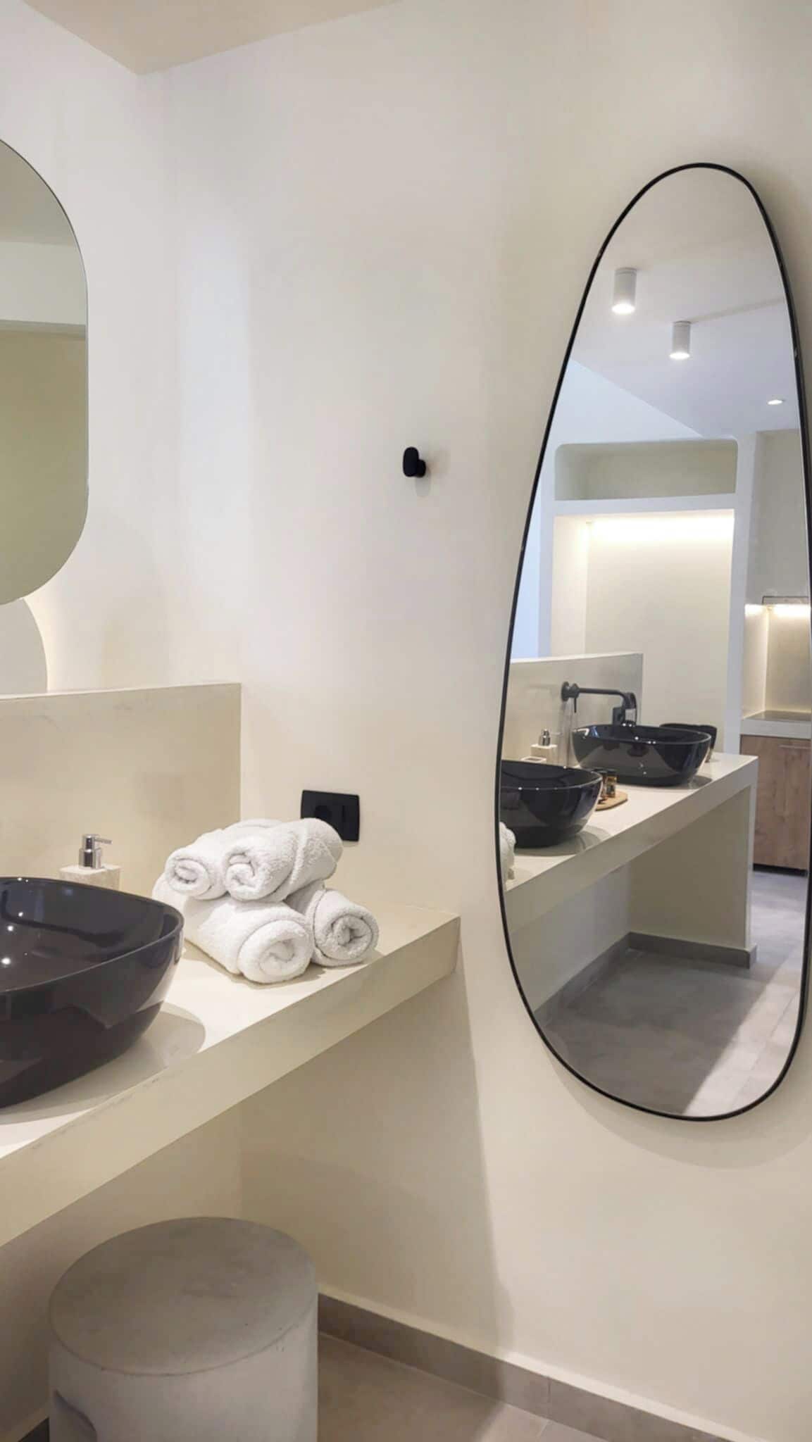 A modern bathroom with a large oval mirror, a round sink, and neatly rolled towels.