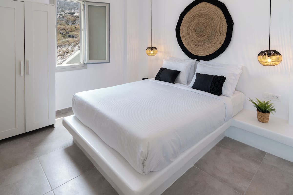 A bedroom with a large bed, a bedside table with a plant, and a view of a mountainous landscape through a window.
