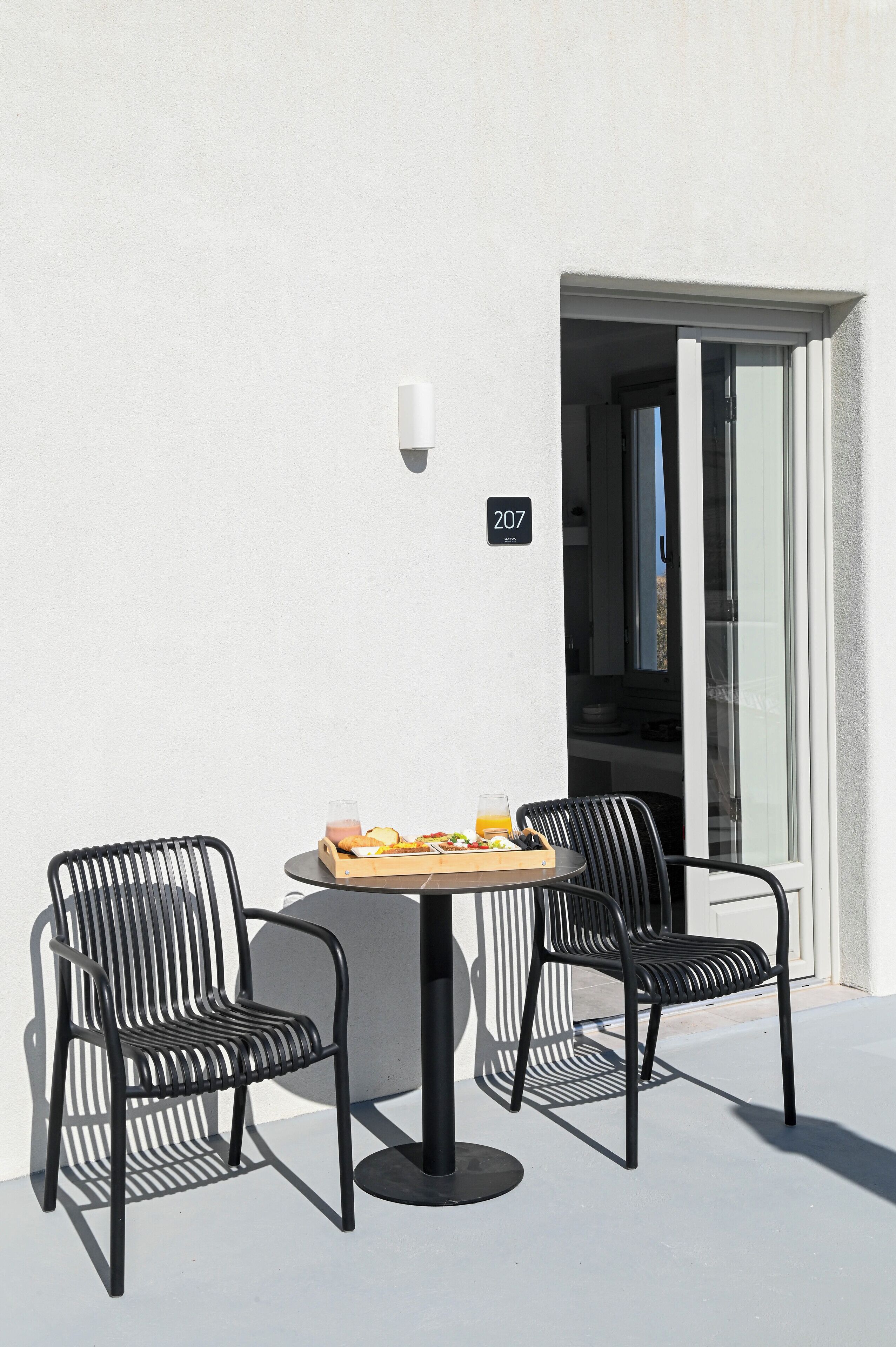 Un petit espace extérieur aménagé avec deux chaises et une table, un panneau indiquant le numéro de l’établissement au-dessus de la porte, et une porte vitrée donnant accès à l’intérieur.