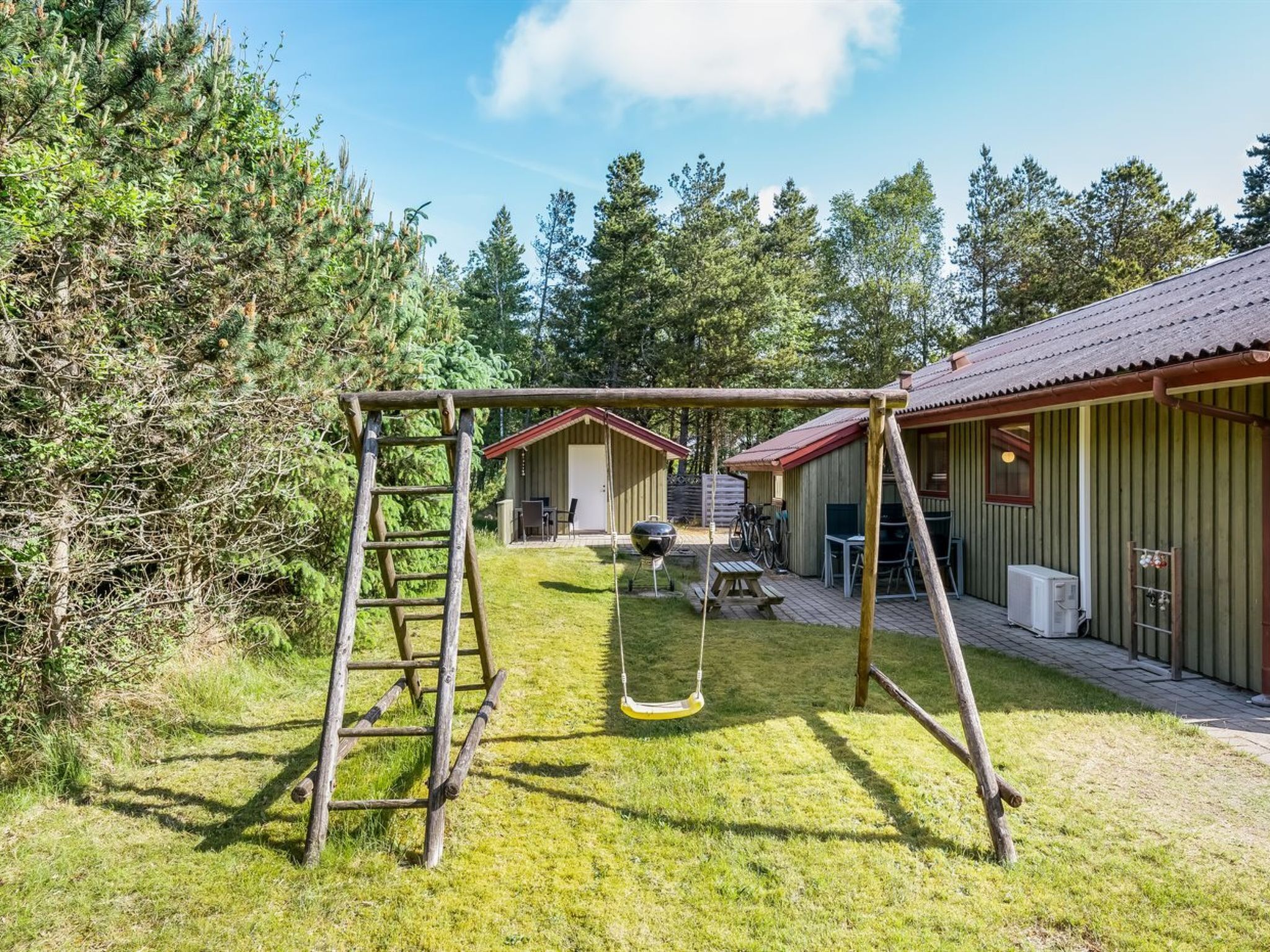 A wooden swing set in a grassy yard with two small sheds in the background.