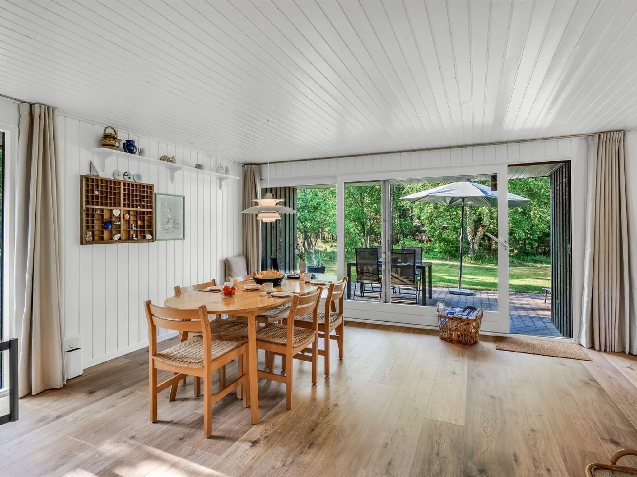 A dining area with a wooden table and chairs, a hanging light fixture, and a view of a garden through large sliding glass doors.