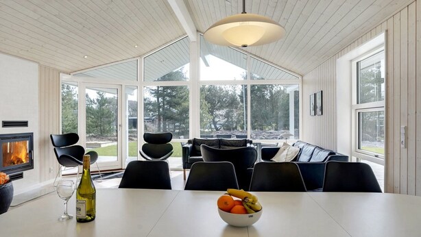 Un espace repas moderne avec une table blanche, des chaises noires, un saladier de fruits, une bouteille de vin et une cheminée.
