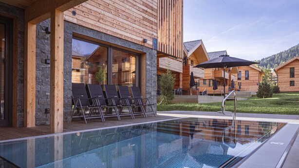 A modern wooden building with a glass infinity pool, outdoor seating, and a clear view of the surrounding mountains.
