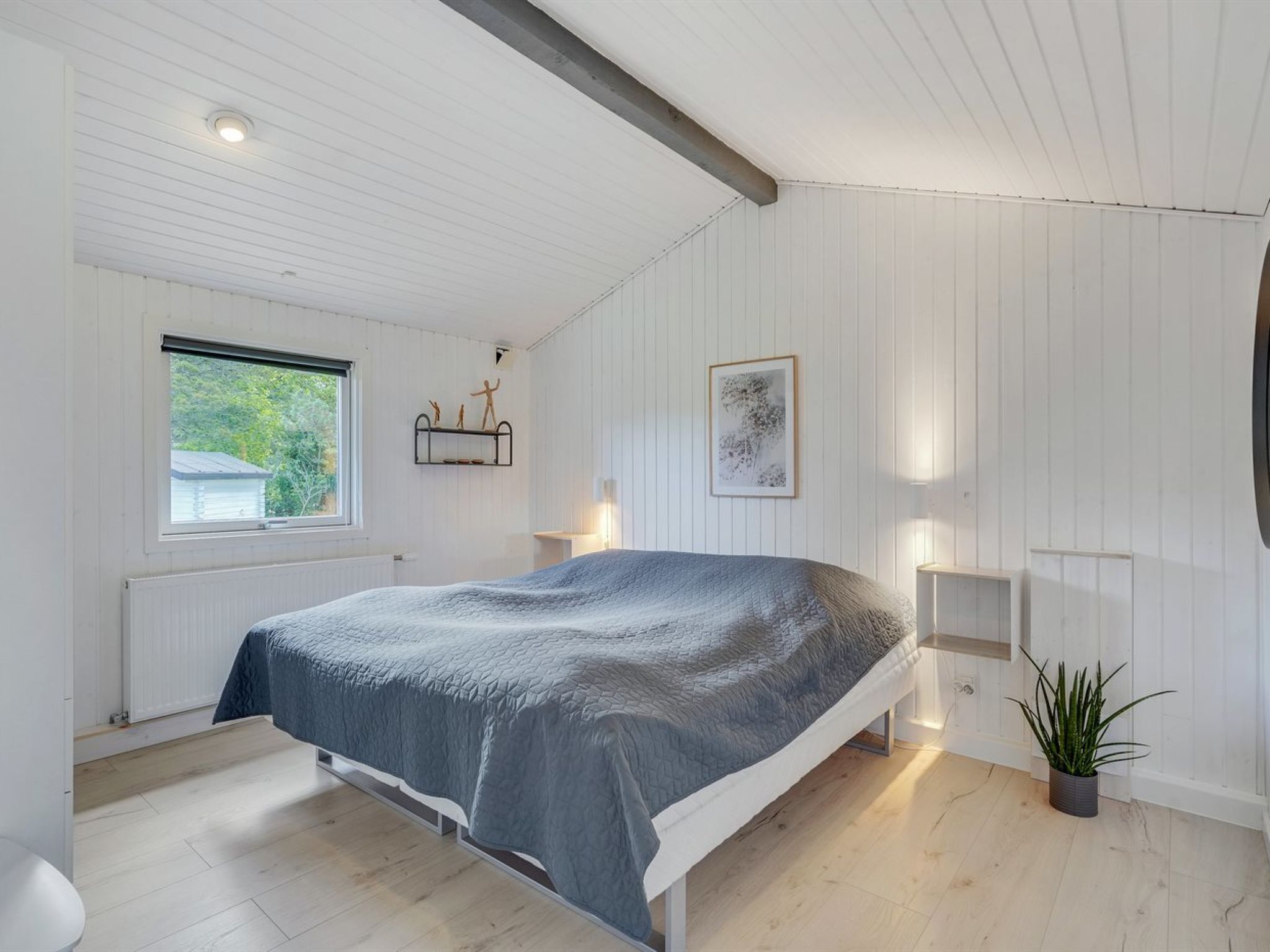 A bedroom with a bed, a window, a radiator, a shelf with decorative items, a framed picture, and a potted plant.