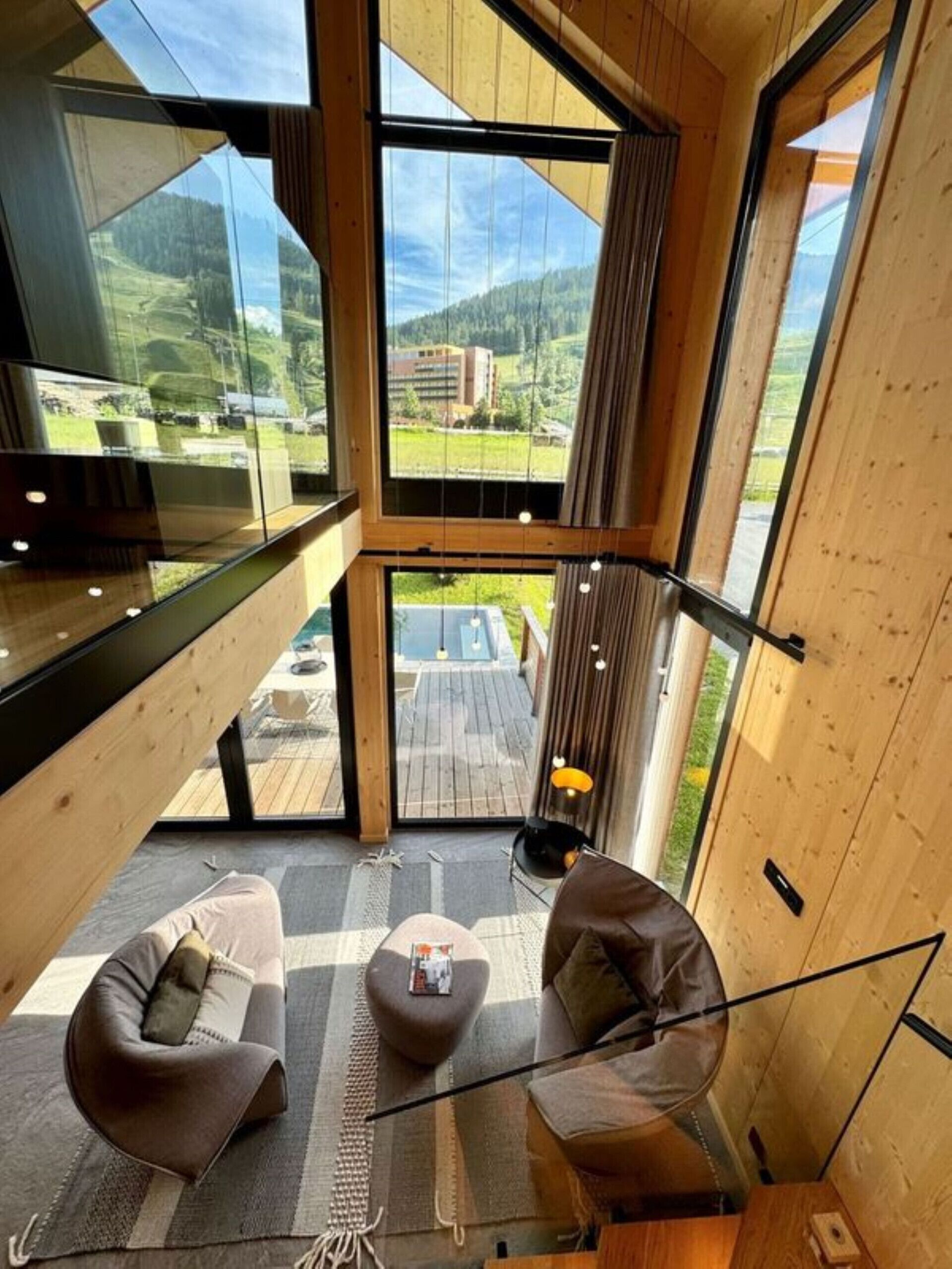 Un intérieur moderne avec un plafond haut, de grandes fenêtres donnant sur un paysage verdoyant et un coin salon confortable avec deux fauteuils et une petite table.
