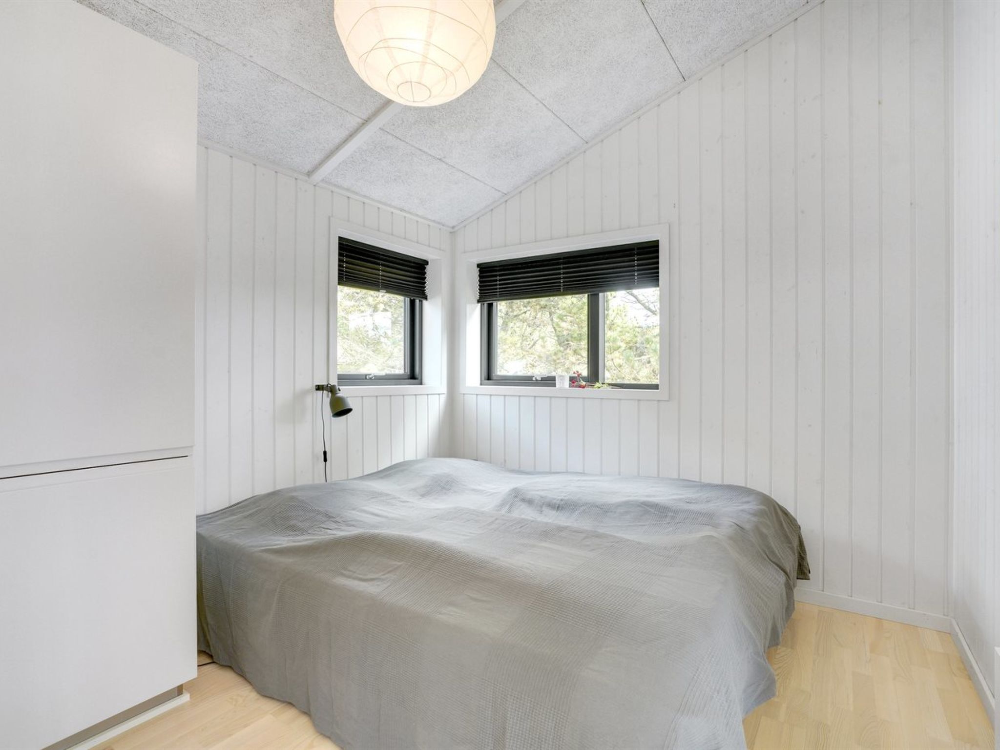 Une chambre à coucher avec un lit, deux fenêtres équipées de stores, un luminaire suspendu et des murs recouverts de panneaux blancs.