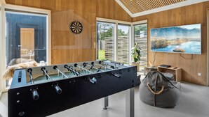 Une chambre moderne équipée d’un baby-foot, d’un jeu de fléchettes et d’un grand écran de télévision diffusant un paysage pittoresque.