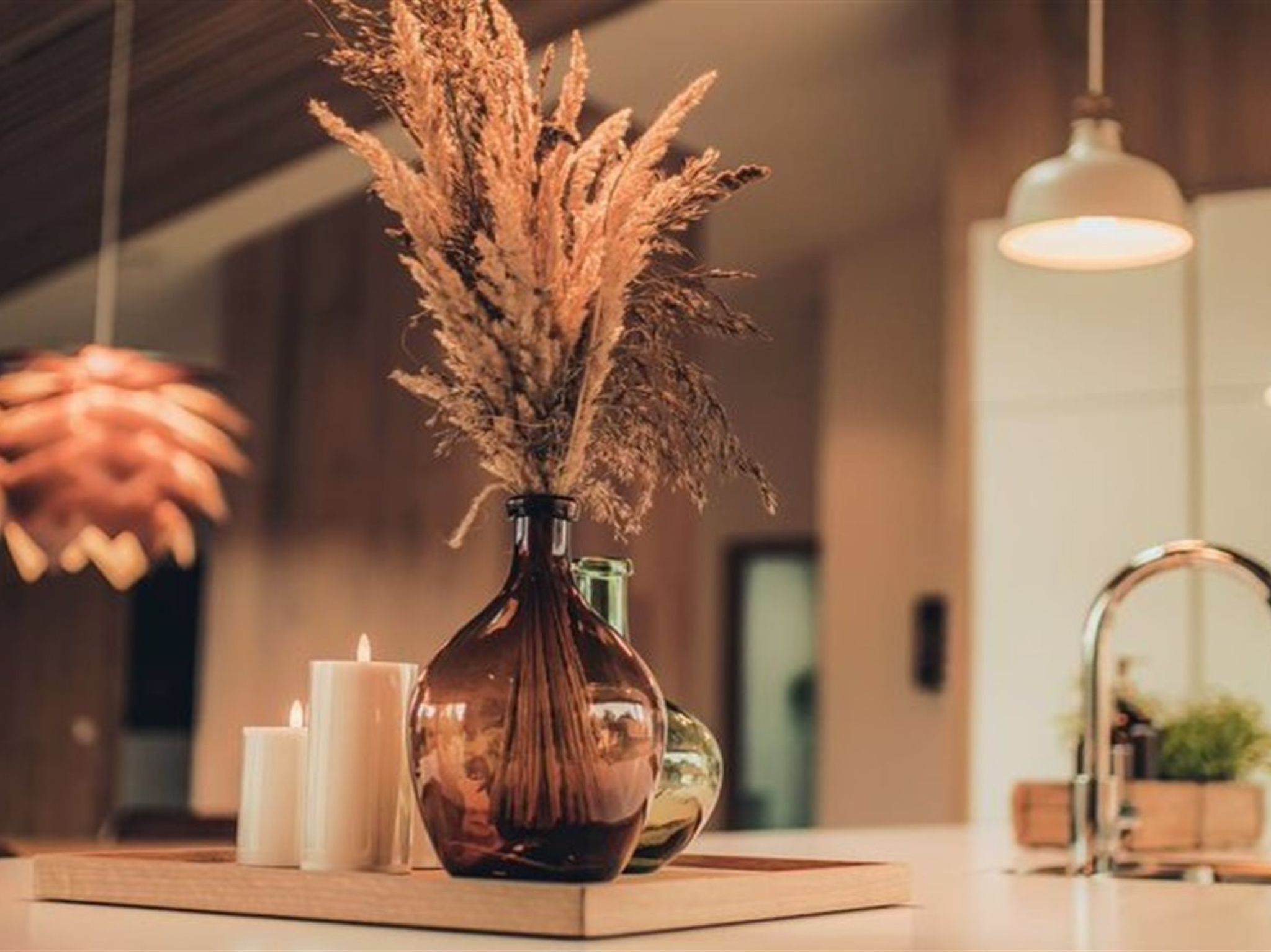 A wooden table with a vase of dried plants, two white candles, and a green bottle.