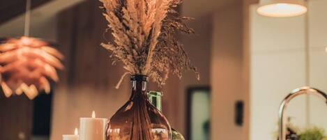 A wooden table with a vase of dried plants, two white candles, and a green bottle.