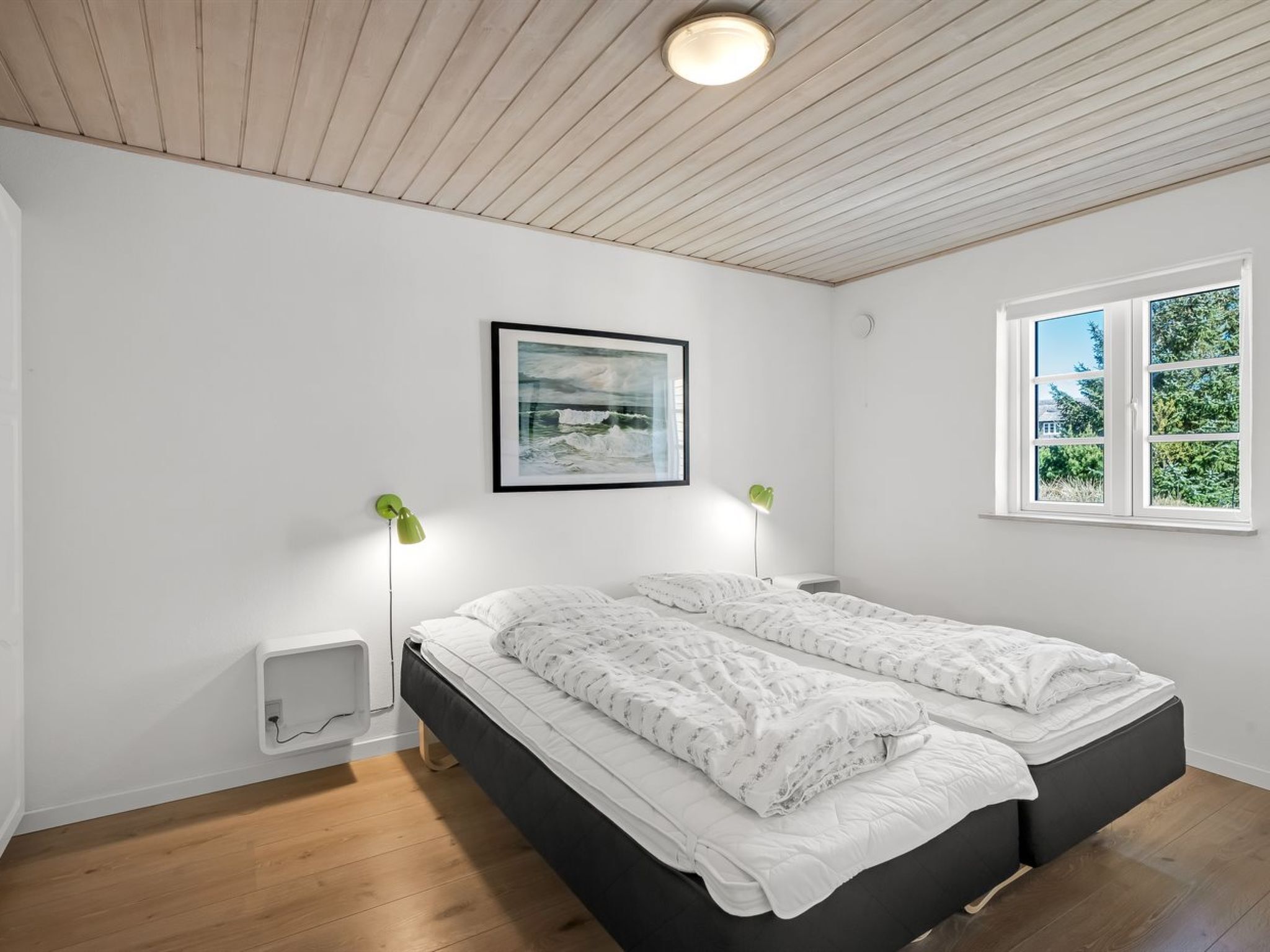 Une chambre avec un plafond en bois, un lit recouvert de linge de lit blanc, deux lampes de chevet, un tableau encadré au mur et une fenêtre donnant sur un paysage verdoyant.