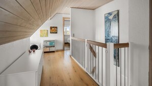 A wooden interior with a sloped ceiling, white walls, and a wooden floor. There is a white railing along the hallway, and artwork is displayed on the walls.