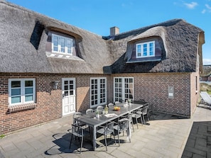 Une maison avec un toit de chaume et des murs en briques, dotée d’une terrasse avec une table et des chaises.