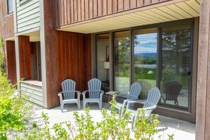 Un bâtiment moderne doté d’un patio couvert, agrémenté de chaises Adirondack blanches et d’une porte en verre donnant sur un paysage pittoresque.