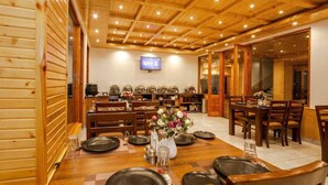A wooden interior with a dining area, tables set with plates and glasses, and a television on the wall. - Mnp Village Venue (Manali)
