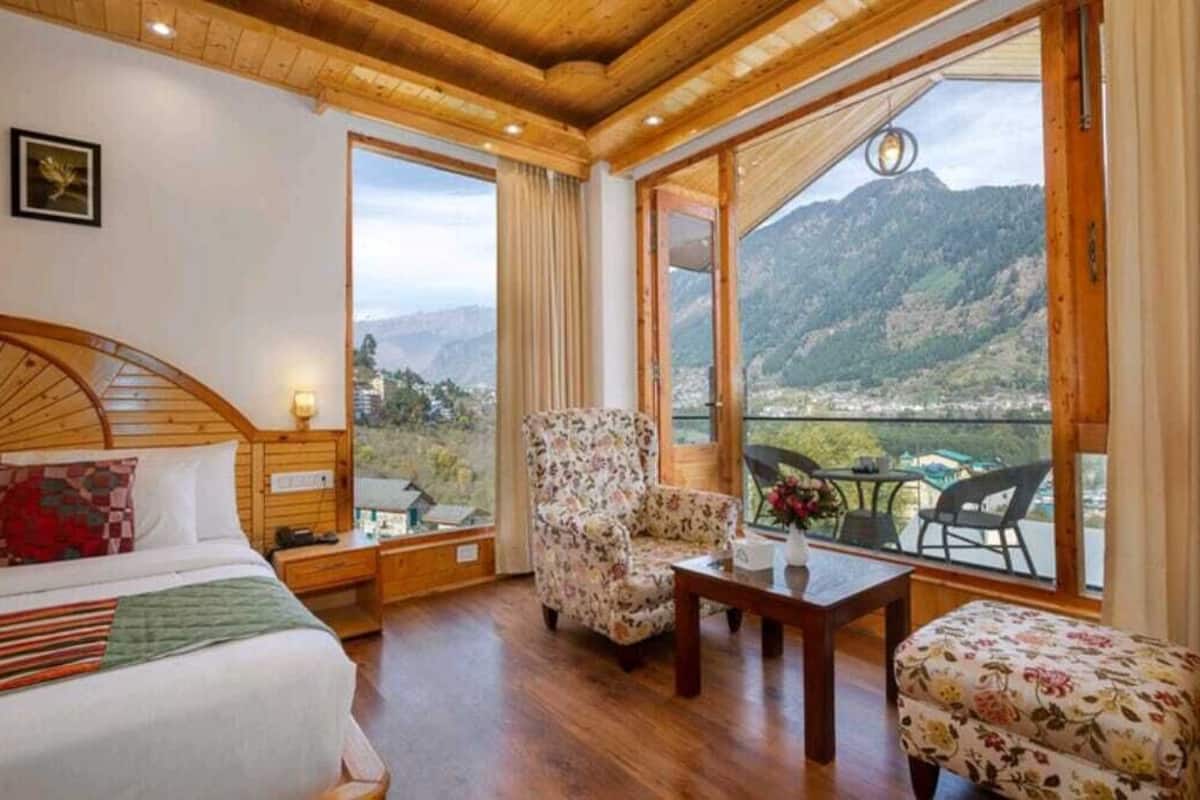 A bedroom with a bed, a chair, a table, and a view of a mountainous landscape through large windows.