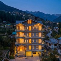A multi-story hotel with a balcony on the second floor, a visible entrance, and a mountainous backdrop.