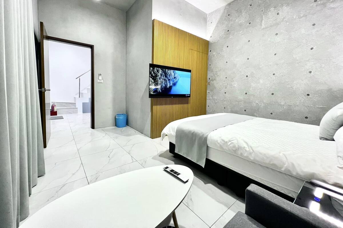 A modern hotel room with a bed, a TV mounted on a wooden panel, a small table, and a bathroom visible through an open door.