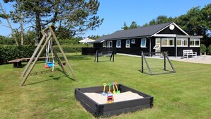 Ein Spielplatz mit Schaukeln, einem Sandkasten mit Schaufeln und einem Spielzeugauto, sowie ein Gebäude mit vielen Fenstern und einer Bank im Hintergrund.