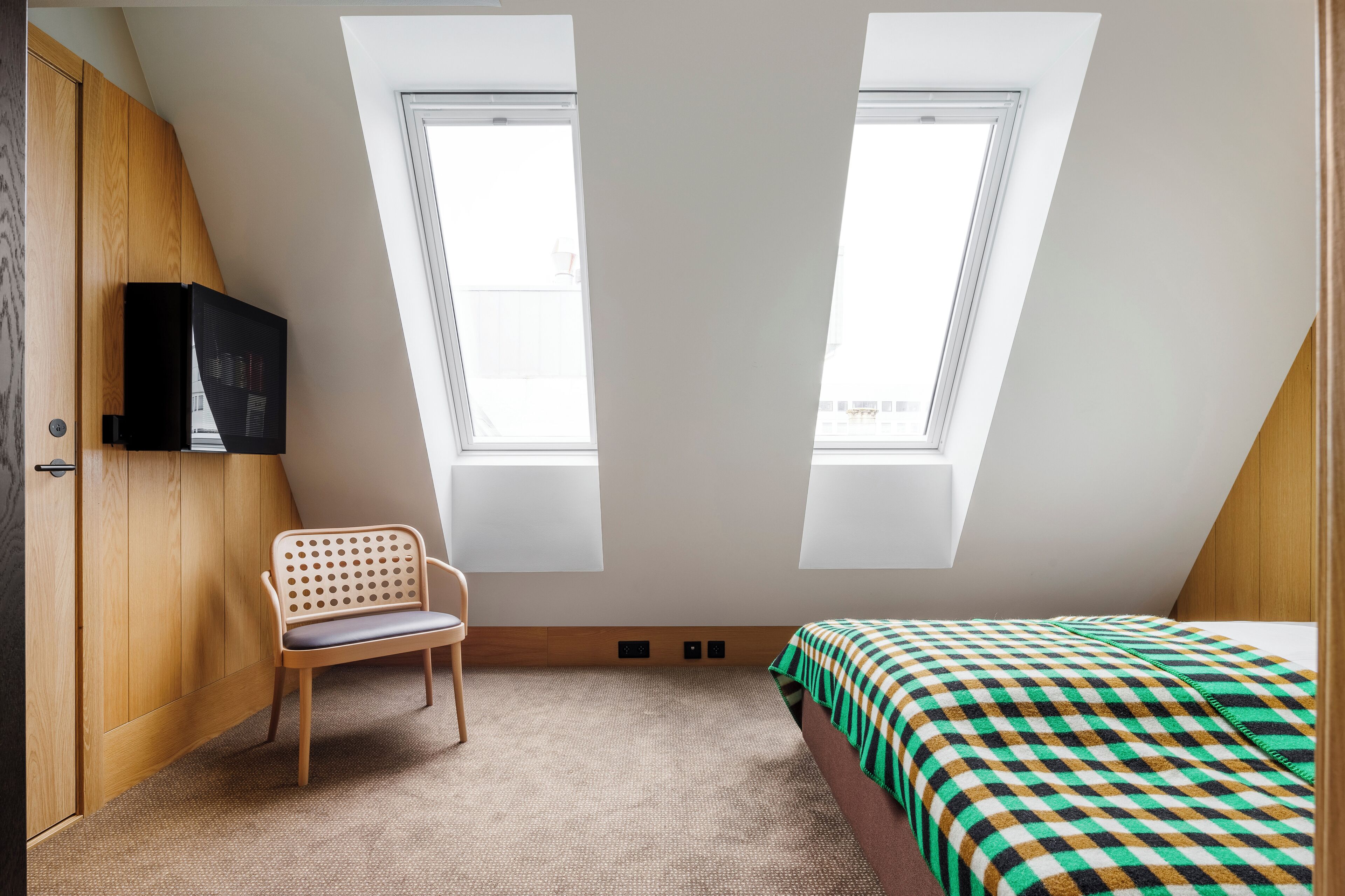 A small, well-lit room with a sloped ceiling, two windows, a bed with a checkered blanket, a wooden chair, and a wall-mounted TV.