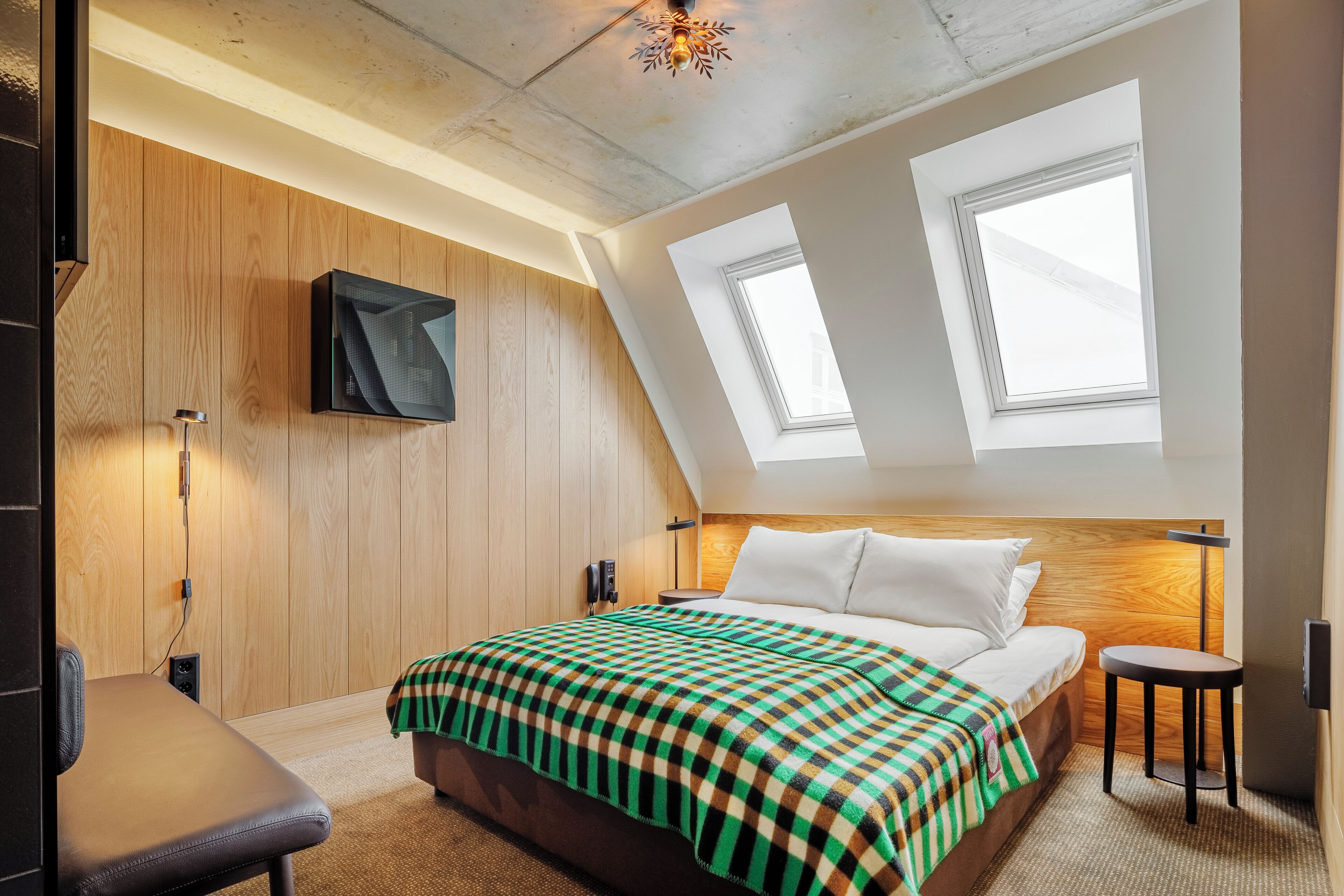 Una camera d'albergo moderna con soffitto a falde, pareti rivestite in legno, un grande letto con coperta a scacchi, una scrivania e una TV a parete.