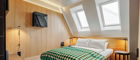 A modern hotel room with a sloped ceiling, wooden panel walls, a large bed with a checkered blanket, a desk, and a TV mounted on the wall.